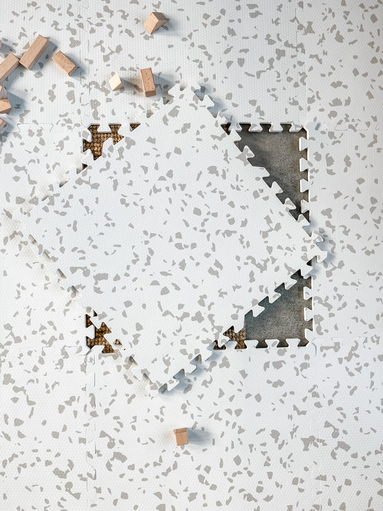 A neutral beige puzzle play mat with a Terrazzo design. One puzzle tile is placed on top of the mat. 