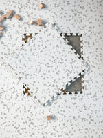 A neutral beige puzzle play mat with a Terrazzo design. One puzzle tile is placed on top of the mat. 